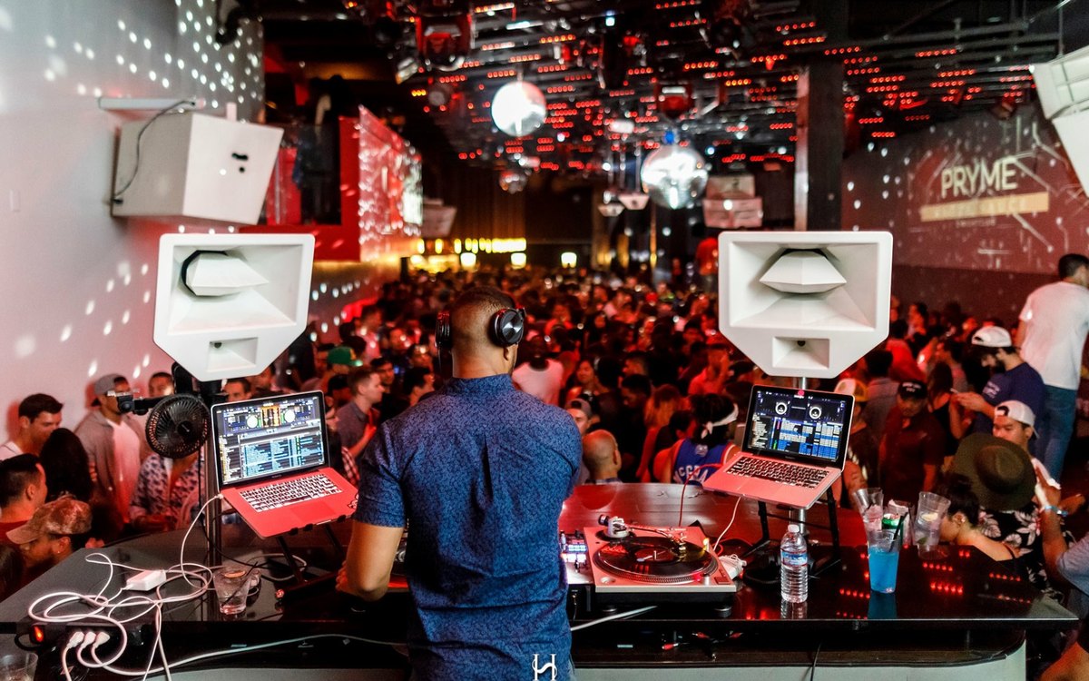 View from behind the decks of deejaypryme playing to a packed red-lit club room with PRYME branding on the screen